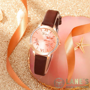 rose gold face watch with 3 dragonflies on it with roman numeral number 12 and 6. with a rose gold trim with  crystals on either side and an elegant brown strap on a beach