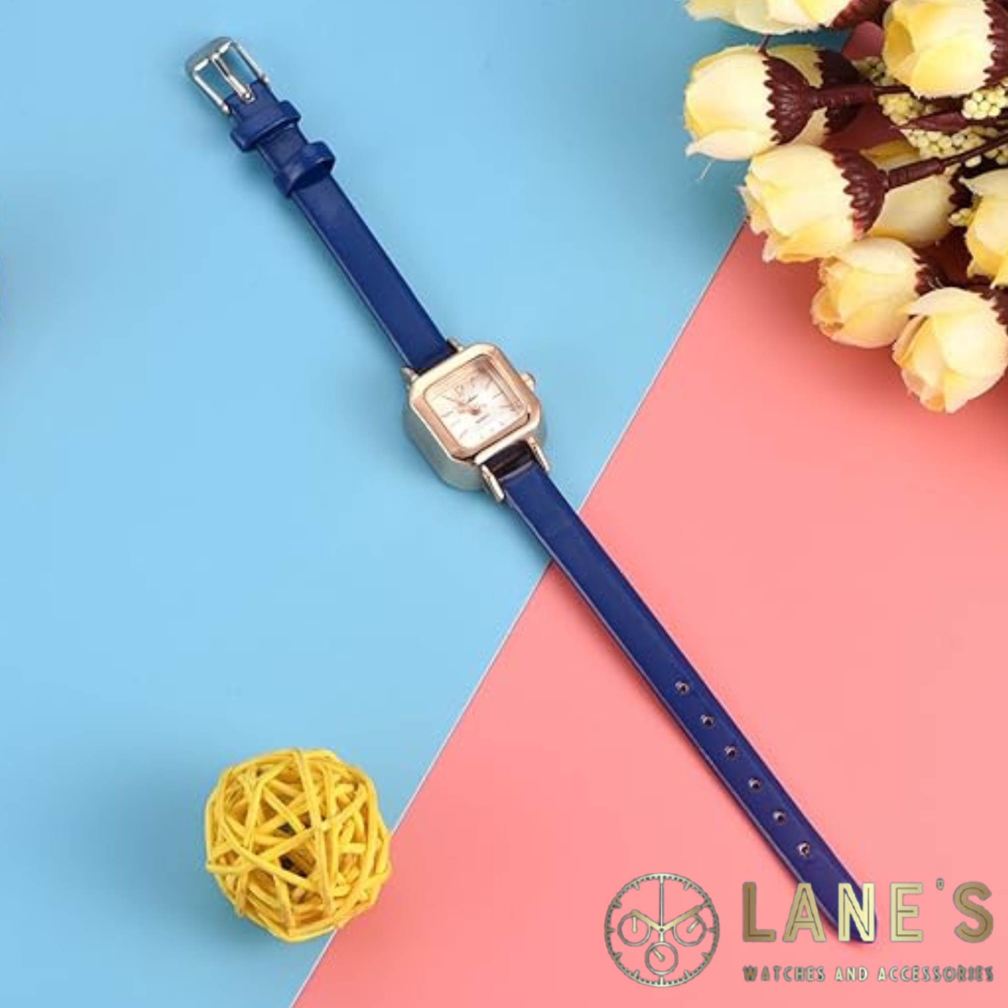 white faced rounded square watch with the numbers 12 and 6 and lines representing the rest of the numbers in a gold colour. with a gold square trim around it and a thin blue strap on a blue and pink background with yellow roses and a yellow rubber band ball