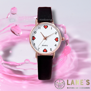 black outlined red hearts at the numbers 12 3 6 and 9 with the rest of the numbers being black on a white faced watch with a gold trim and a black textured strap on a pink background