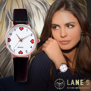 black outlined red hearts at the numbers 12 3 6 and 9 with the rest of the numbers being black on a white faced watch with a gold trim and a black textured strap on a woman's wrist