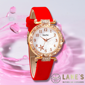 white faced watch with gold numbers and a gold trim with crystals and a red strap on a pink background